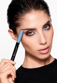 Niebieska kredka do oczu przyłożona do brwi, z błyszczącą czarną obudową oznaczoną napisem "Made to Last". Modelka ma teksturowaną, gładką fryzurę i subtelny makijaż.