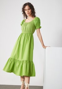 Grünes Baumwoll-Midikleid mit kurzen Puffärmeln, quadratischem Ausschnitt, gerafftem Oberteil und rüschengesäumtem Saum. Glatte Textur mit fließender Silhouette.