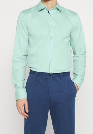 Mann trägt ein hellgrünes langärmeliges Hemd mit Knöpfen, in blaue Hosen gesteckt, eine Hand in der Hosentasche, vor einfarbigem Hintergrund.