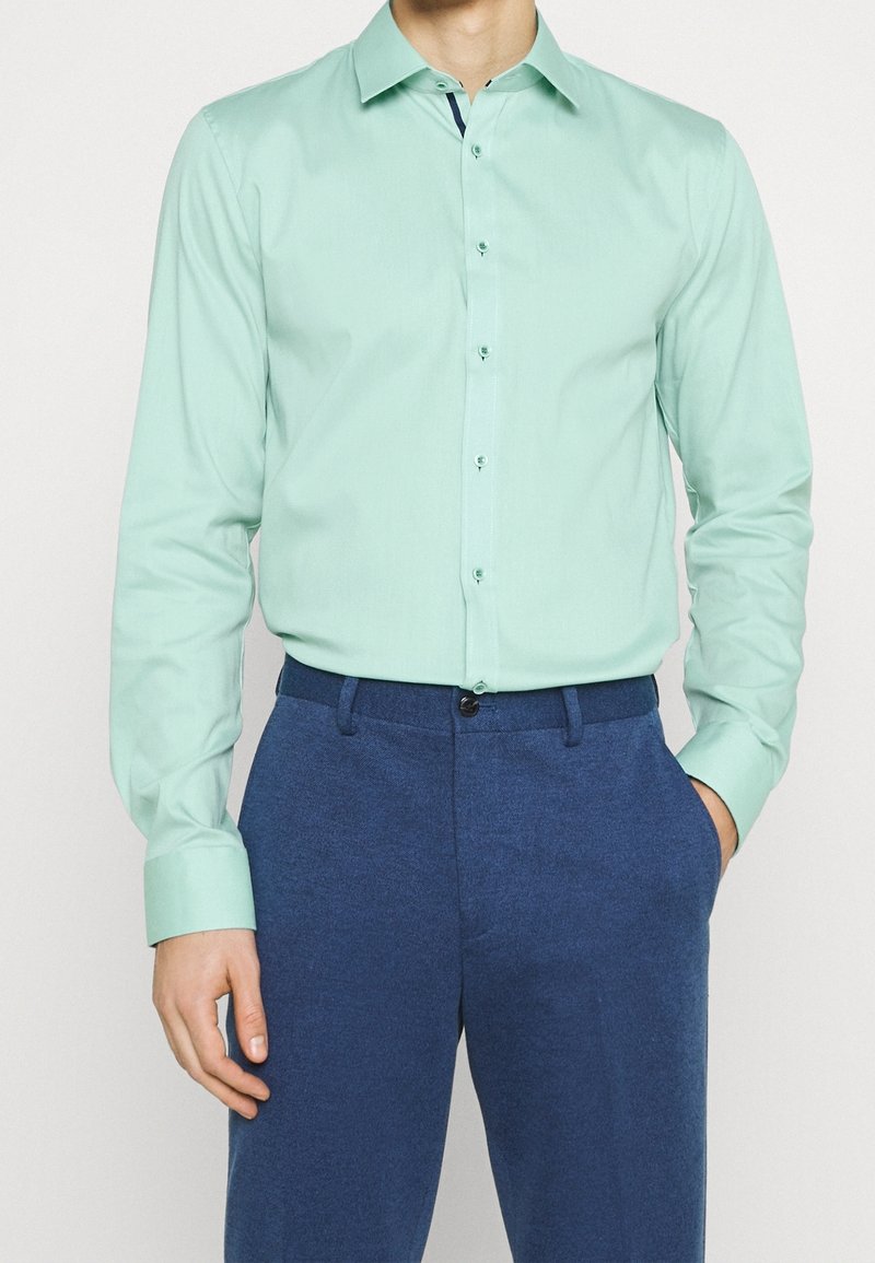 Homme portant une chemise boutonnée à manches longues vert clair rentrée dans un pantalon bleu, une main dans la poche, sur fond uni.