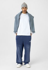 Blauwe wijde sweatpants, wit T-shirt met ronde hals, en een grijze sweatshirt over de schouders gedrapeerd. Een grijze gebreide beanie en witte sneakers maken de look af.