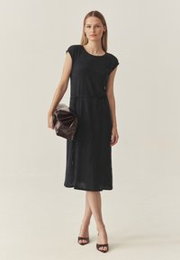 Crna haljina do koljena s kratkim rukavima, teksturirana tkanina i strukirana. Dodaci uključuju tamnosmeđu kožnu torbu i sandale s petom.