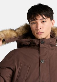 Brun parkas med huva fodrad med fuskpäls, knäppning framtill och sidofickor. Tyget har en jämn textur med matt yta.