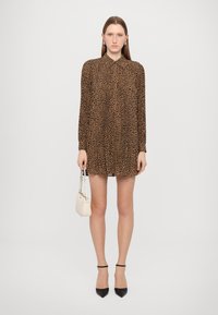 Långärmad klänning med leopardtryck i brunt och svart, med krage och plisserad design, i kombination med en liten vit handväska och svarta klackskor.