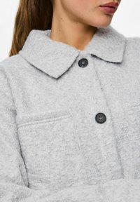 Veste boucle grise avec un col évasé, fermeture à boutons noirs et une petite poche poitrine. Tissu doux et texturé avec un aspect duveteux.
