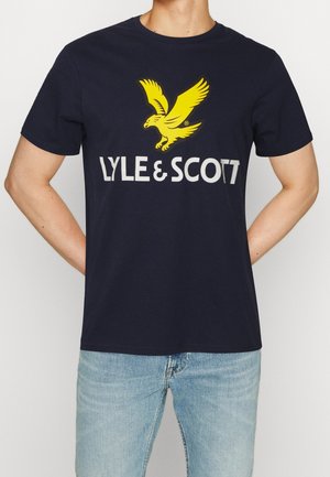 Personne portant un t-shirt bleu marine avec un logo d'aigle jaune et le texte blanc LYLE & SCOTT, associé à un jean bleu clair.