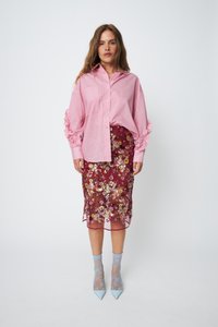 Pink striped shirt with oversized ruffle sleeves, paired with a burgundy floral embroidered sheer skirt. Light blue patterned socks and pointed heels.