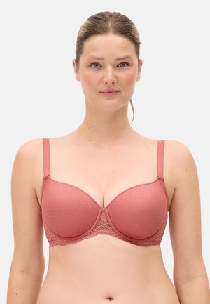 Mujer de piel clara y cabello recogido que lleva un sujetador acolchado color rosa polvoriento con ribete de encaje frente a un fondo liso.