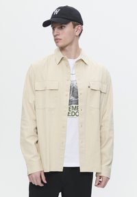 Beige button-up shirt with two front pockets, worn over a white graphic t-shirt featuring green text. Black cap with logo. Black pants.