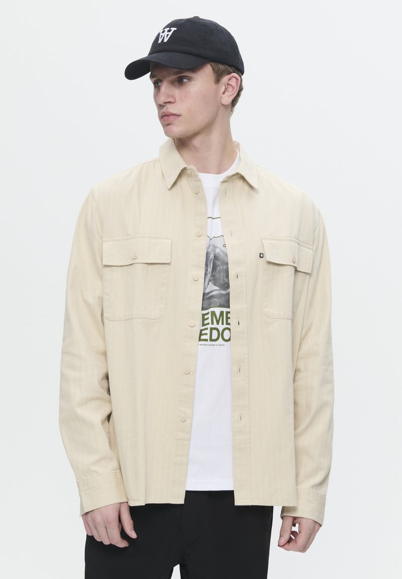 Beige button-up shirt with two front pockets, worn over a white graphic t-shirt featuring green text. Black cap with logo. Black pants.