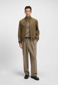 Olijfgroene bomberjack met rits, gecombineerd met een beige coltrui, beige los vallende broek en zwarte schoenen. Gladde textuur, modern ontwerp.