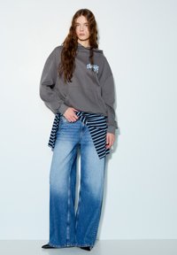 Sivi veliki hoodić s plavim logotipom, uparen s širokim trapericama blijede boje. Tkanina s prugama vezana oko struka dodaje detalj.