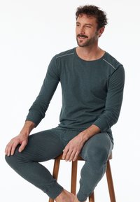 Maglietta a maniche lunghe di un blu verde scuro con pantaloni abbinati in materiale morbido. Presenta cuciture bianche sottili sulle cuciture delle spalle e lungo i lati dei pantaloni.