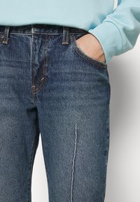 Jean denim bleu foncé avec surpiqûres jaunes, design à cinq poches et coupe droite. Le tissu présente une finition légèrement texturée.