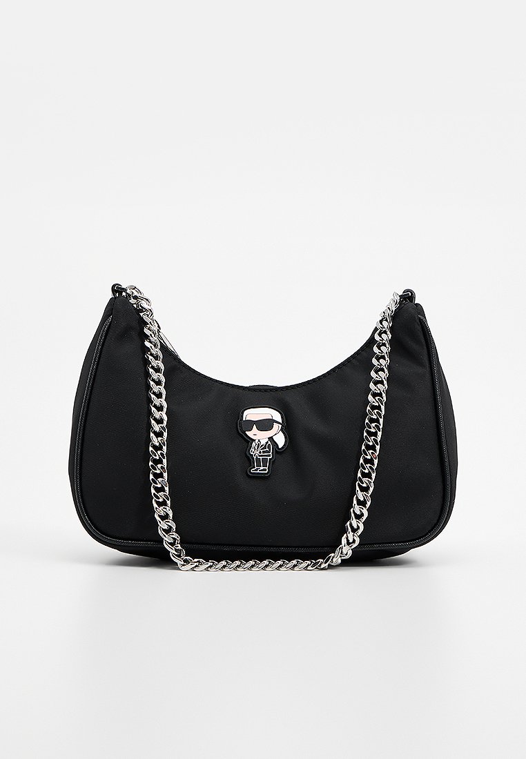 Sac en tissu noir avec une forme arrondie, une bandoulière en chaîne argentée et un détail de broche en forme de cartoon à l'avant. Texture lisse, design minimaliste.