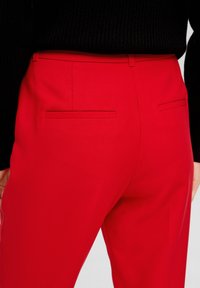 Czerwone spodnie o dopasowanym kroju wykonane z gładkiego materiału, slim fit, z dwoma tylnymi kieszeniami i poziomymi szwami. Noszone z czarną bluzką w prążki.