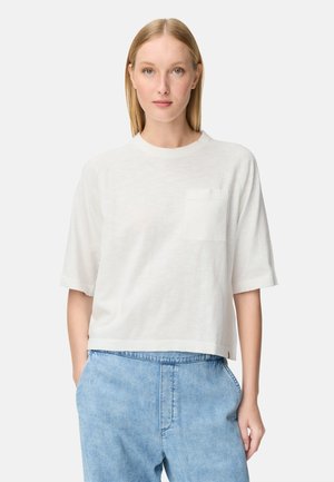 Blonde Frau steht frontal, trägt ein weißes kurzärmeliges Oberteil mit Brusttasche und hellblaue, hoch taillierte Jeans.
