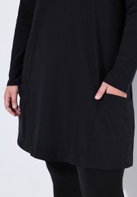 Schwarze Tunika mit langen Ärmeln, ausgestattet mit Seitentaschen. Hergestellt aus weichem, glattem Stoff mit lockerer Passform, die oberhalb des Knies endet.