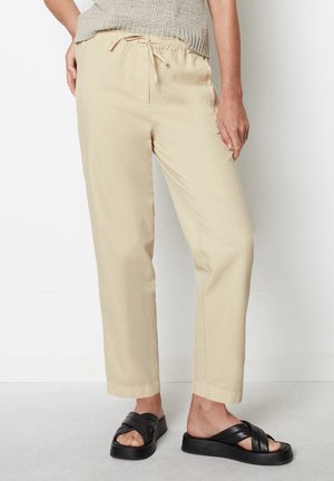 Beige broek met trekkoord, gedragen met zwarte platformsandalen en een licht gebreid mouwloos topje, staand tegen een witte achtergrond.