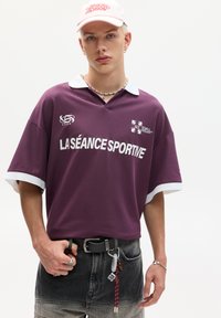 Purple short-sleeve polo shirt with white collar, featuring "LA SÉANCE SPORTIVE" text. Worn with black belt and distressed grey shorts.
