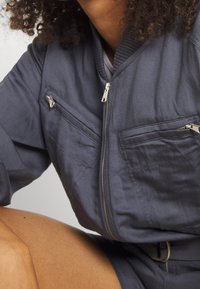 Chaqueta bomber de color gris oscuro, confeccionada en un tejido suave, con bolsillos de pecho con cremallera, cuello de canalé y detalles de costura visibles.