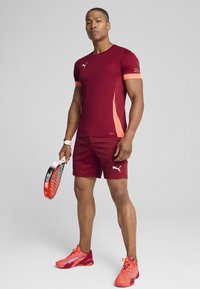 Herre athletic outfit i bordeaux med orange detaljer; tætsiddende kortærmet skjorte og shorts, parret med klare røde og pink sneakers.