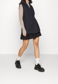 Námořnická vesta s dvojitým běžcem a širokými klopami, spárovaná s béžovým topem s dlouhým rukávem, černými šortkami a robustními černými šněrovacími botami.