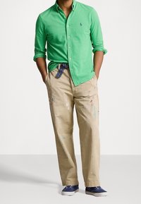 Man med grön knappad skjorta, beige byxor med färgfläckar och randigt bälte, samt blå sneakers. Han har händerna i fickorna och står framför en enfärgad bakgrund.