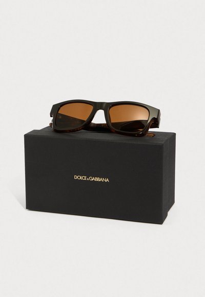 Melna rāma saulesbrilles ar brūnām lēcām, kas atpūšas uz melnas Dolce & Gabbana kastes ar zelta logo uz baltas fona.