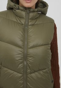 Doudoune sans manches vert olive avec capuche, dotée d'une fermeture éclair, d'un tissu texturé et d'accents de boutons sur le côté. Les manches en tricot marron contrastent avec la doudoune.