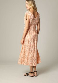 Femme portant une robe midi sans manches orange et blanche à motifs avec des bretelles à nouer, sandales noires, se tenant de dos, regardant à gauche.
