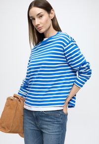 Modrá a bielo pruhovaná košeľa s dlhým rukávom, s okrúhlym výstrihom, voľného strihu, so svetlomodrými rifľami a hnedou semišovou kabelkou.