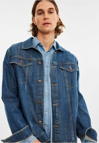 Tummansininen farkkutakki, jossa on klassinen kaulus, etupainikkeet ja kaksi rintataskua, kerrostettuna vaaleansinisellä nappikauluspaidalla.