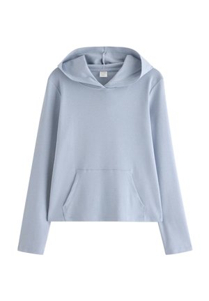 Jeune femme en sweat à capuche bleu clair et pantalon assorti, debout, souriante, les mains dans la poche avant, sur un fond blanc uni.