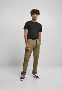 Camiseta negra de manga corta, pantalones color caqui claro con cinturilla elástica y cinturón negro, zapatillas blancas y negras, de pie sobre un fondo liso.