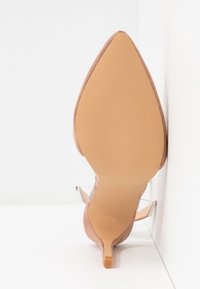 Chaussure à talon haut beige à bout pointu avec lanières transparentes à la cheville, vue de la semelle vers le haut sur un fond blanc.