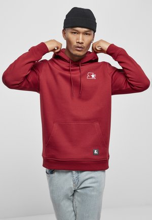 Rød hettegenser laget av mykt stoff, med kengurulomme, snorhette og en liten grafisk logo på brystet. Brukt med lyse jeans.