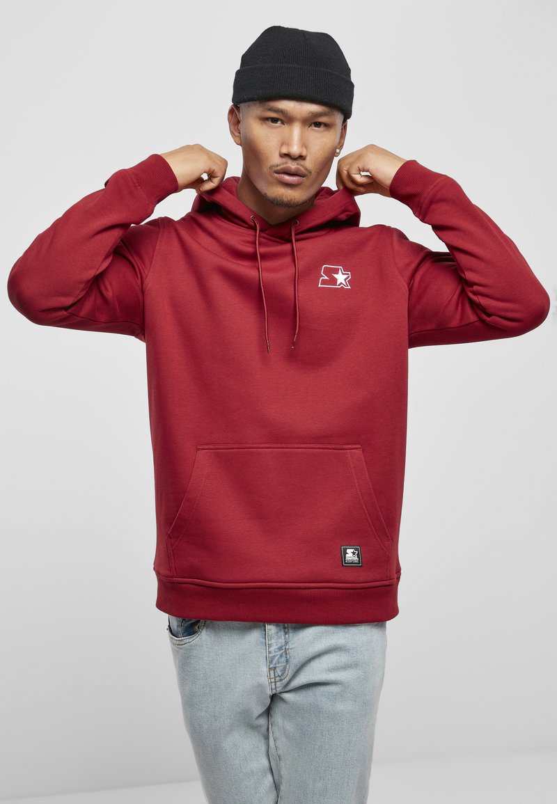 Rød hettegenser laget av mykt stoff, med kengurulomme, snorhette og en liten grafisk logo på brystet. Brukt med lyse jeans.