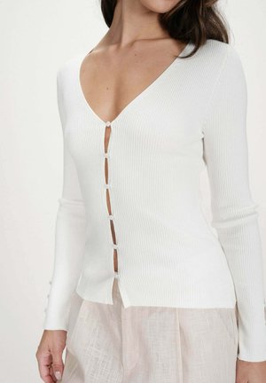 Femme portant un cardigan blanc côtelé à manches longues avec de petits boutons et un pantalon beige léger, montrée du cou aux hanches.