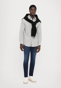 Hvit mønstret skjorte med en svart genser drapert over skuldrene, blå slim-fit jeans og hvite sneakers med en beige såle.