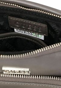 Borsa in pelle grigia con chiusura a zip. L'interno presenta una fodera in tessuto nero con il logo 'GUESS' ripetuto. Etichetta in pelle visibile all'interno.