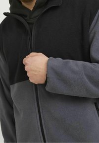 Giacca in pile con blocchi di colore grigi e neri, zip completa, colletto alto e tasche laterali con zip. Tessuto morbido, progettata per il calore.