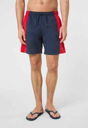 Hombre con pantalones cortos de baño azul marino y rojo con cintura de cordón y chanclas azul marino, de pie frente a un fondo liso.