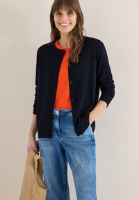 Jeune femme souriante, portant un cardigan noir boutonné sur une chemise orange et un jean bleu, tenant une veste beige dans une main.