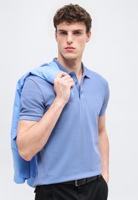 Νεαρός άνδρας με ανοιχτό μπλε πόλο πουκάμισο κρατάει ταιριαστό σακάκι στον ώμο, στέκεται μπροστά σε απλό φωτεινό φόντο.