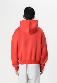 Roter Kapuzenpullover mit lockerer Passform, geripptem Saum und Bündchen sowie strukturiertem Stoff. Getragen mit grauen Jogginghosen.