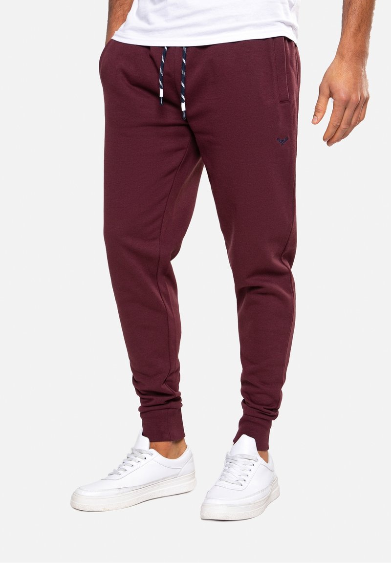 Bordó tepláky s elastickými manžetami na kotnících, bočními kapsami a stahovacími šňůrkami v pase. Kombinovány s bílými teniskami. Jednoduchý design, měkká textura.