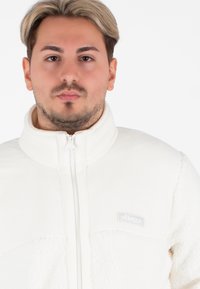 Witte fleecejack met een hoge kraag en rits, met een mix van gladde en gestructureerde stof, en een klein logo op de borst.