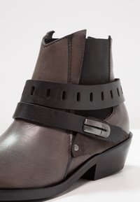 Brązowe skórzane botki za kostkę o matowym wykończeniu, z dekoracyjnym czarnym paskiem i klamrą, elastycznymi bocznymi panelami oraz niskim obcasem.