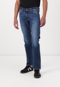 Mørkeblå denimjeans med rette ben, synlige sømmer og fem lommer. Brukt med svarte sneakers med hvite detaljer.